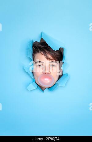 Boy blowing bubble with head through hole in blue paper background. Stock Photo