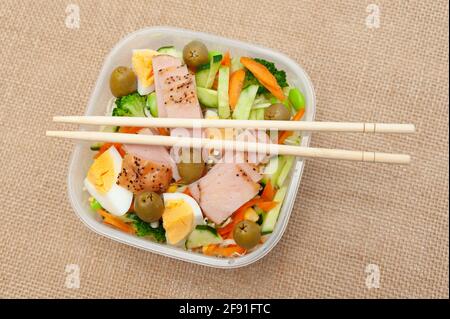 Healthy chicken salad with vegetables in plastic container with chopstick. Isolated on jute background. Top view. Stock Photo