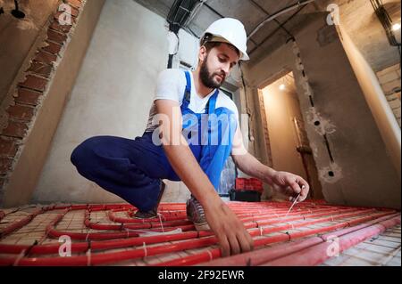 Horizontal, low angle view portrait of young plumber wearing blue overalls and white helmet, tying up red tubes on the floor heating system in new unfinished apartment Stock Photo