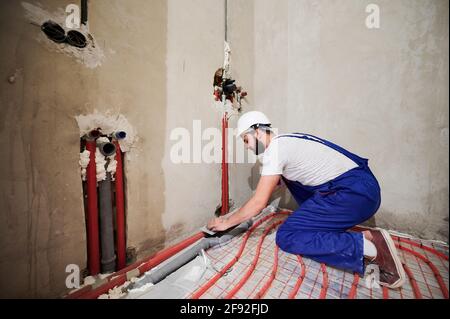Wide angle view on young worker standing on his knees in blue overalls and helmet installing underfloor heating system and plumbing system. Empty room with many pipes. Construction concept Stock Photo