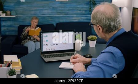 Tired elderly entrepreneur checking graphics from laptop writing on notebook working from business office overtime. Busy employee using modern technology network wireless reading typing, searching Stock Photo