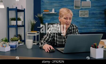 Old middle aged businesswoman working at laptop typing financial data sitting at desk using modern technology while retired husband sitting on couch. Serious lady analyzing and managing domestic bills Stock Photo