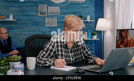 Focused stylish mature old woman remote working from home distance office on laptop taking notes. Middle aged business lady using computer watching webinar sitting on chair writing in notebook. Stock Photo