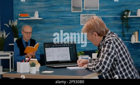 Senior woman architect analysing digital prototype with plans from laptop working from home, taking notes on notebook. Elderly designer using cad software to create a 3D concept of buildings Stock Photo