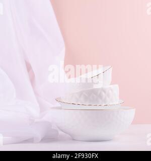 White ceramic dishes pile with thin gold border with airy silk curtain on white wood table in delicate pastel pink interior, square. Stock Photo