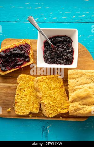 Cornbread is bread containing cornmeal, corn flour. Beautiful yellow sliced cornbread loaf with blueberry jam in bowl. Blue wood background. Flat lay Stock Photo