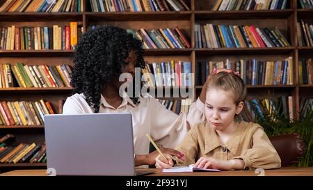 Black lady hugs sad adopted blonde girl sitting at brown table with grey laptop and writing in copybooks against racks with books at online lesson Stock Photo