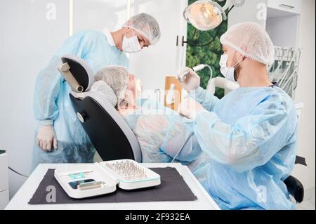 Horizontal snapshot in dentist office of dentist and his assistant during surgery performance. People wearing disposable closing , gloves, caps and masks. Surgical taper kit on the table. Stock Photo