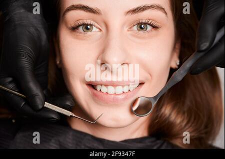 Close up portrait of beautiful young smiling woman lying in dental chair. Dentist hands in sterile gloves using dental explorer and mirror while examining female patient teeth. Concept of dentistry. Stock Photo