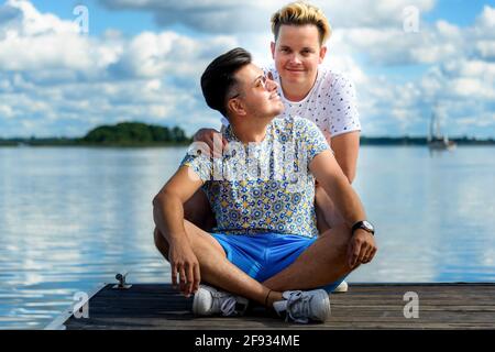 happy smiling gay couple sitting on a wooden pier against lake and blue sky Stock Photo