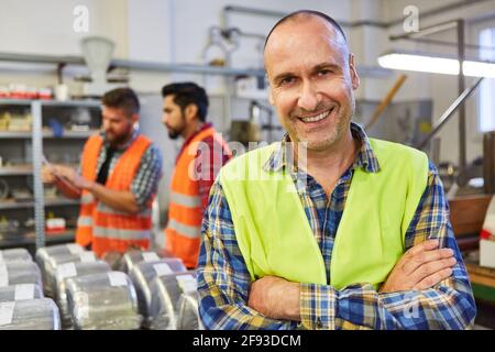 Happy skilled worker stands in front of the warehouse of a metal factory with a team in the background Stock Photo