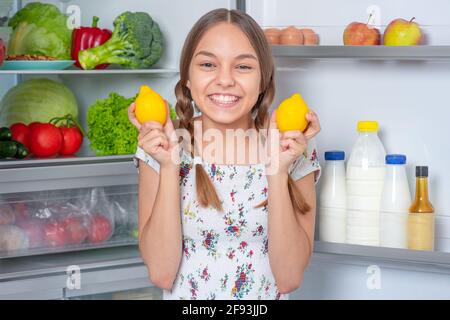 Beautiful young happy teen girl holds fresh yellow lemons while standing near open fridge in kitchen at home Stock Photo