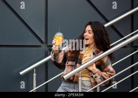 Young stylish woman with juice cup standing over gray background. Portrait of a smiling woman with curly hair. Stock Photo