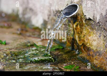 water source flows out of a metal pipe from an old wall made of large old stones in the sunlight with reflections on a stone overgrown with moss Stock Photo