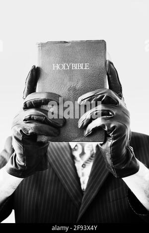 Man wearing black leather gloves and pinstripe suit holding up bible to cover his face on white background.  Concept of intrigue, mystery, curiosity Stock Photo