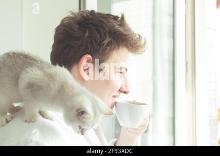 Young caucasian man is drinking coffee near the window. He is holding a cat and smile. Early morning, sunshine. Stock Photo