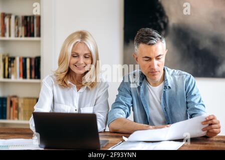 Two focused successful and confident business partners in stylish clothes are sitting at the workplace, discussing, working on a new project, carefully reviewing and checking documents, use laptop Stock Photo