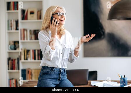 Happy friendly busy mature caucasian woman, in stylish wear and glasses, working at office, standing near desk, talking to employee or client on the cell phone, gesturing with hand, smiling Stock Photo