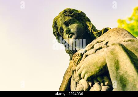 Weathered statue of an Angel with cracked dirty face (with a spider) covered with moss at old cemetery - cross processed image Stock Photo