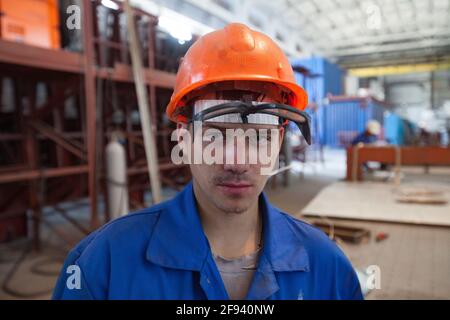 Young mechanic worker in orange hardhat and protective glasses. machine production plant background. Stock Photo