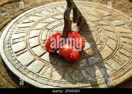 Small red Easter eggs hidden in a spring garden ready to be found. Brightly coloured foil covered chocolate eggs perched on a sundial. Stock Photo