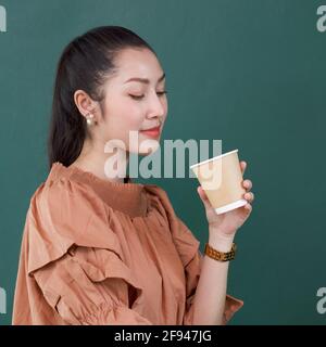Young asian woman in casual dress looking at coffee drink in paper cup. Portrait on green background with studio light.