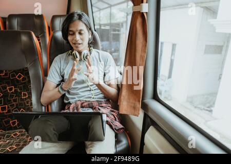 a man with hand gestures and headphones uses a laptop while chatting using a virtual meeting while sitting near a window Stock Photo