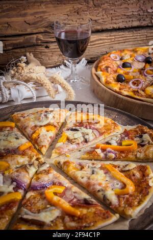 Tasty homemade pizza with salami and vegetables and a tuna pizza in the background on a rustic wooden table served with red wine Stock Photo