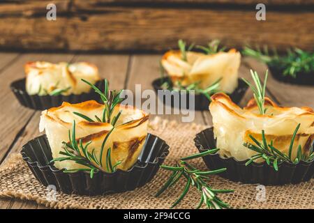 Baked crispy potato roses with rosemary in black tartlet forms on a rustic wooden table Stock Photo
