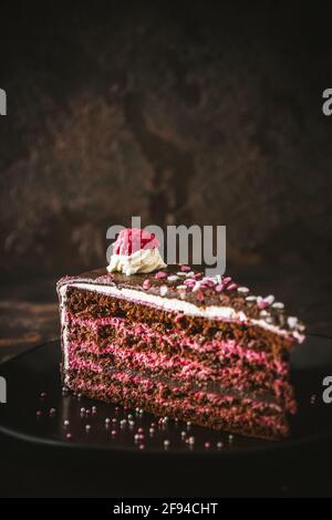 Piece of raspberry chocolate torte on a black plate on dark brown background, vertical with copy space