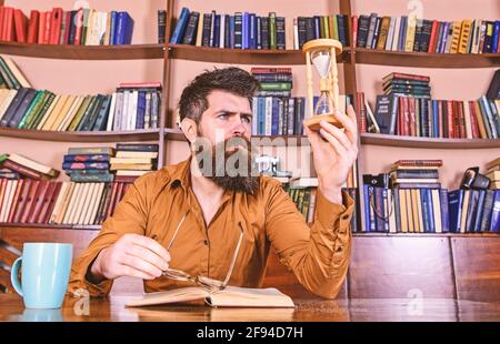 Man on busy face reading book, bookshelves on background. Lesson and time concept. Scientist with eyeglasses sits at table and looks at hourglass Stock Photo