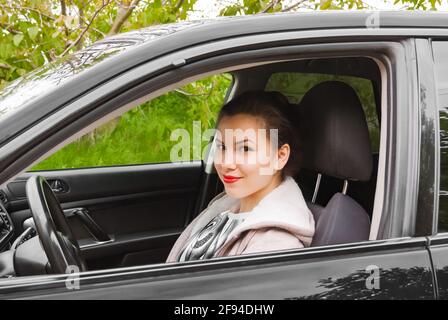 A young woman is sitting in a car smiling and looking at the camera through the window opening Stock Photo