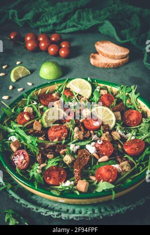 Bowl of fresh vegetable salad: arugula, fresh and dried cherry tomatoes, pine nuts and cheese on a green background, vertical Stock Photo