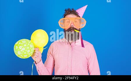 Celebration concept. Man with beard and mustache on calm face blows into party horn, blue background. Hipster in giant sunglasses celebrating birthday Stock Photo