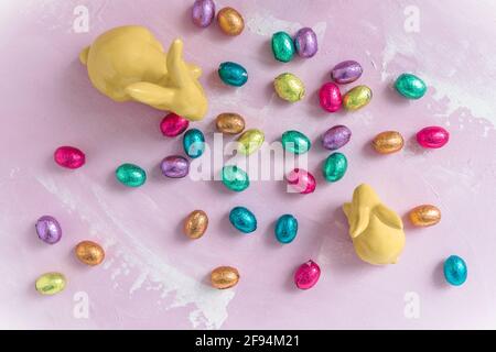 Two yellow porcellain easter bunnies with colorful chocolate easter eggs on pinkish background, easter decoration, top view Stock Photo