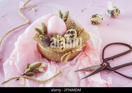 Scene of handmade Easter decoration: jute fabric, pink Easter egg, natural quail eggs, feathers and branches on pinkish background Stock Photo