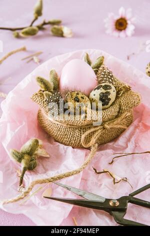 Scene of handmade Easter decoration: jute fabric, pink Easter egg, natural quail eggs, feathers and branches on pinkish background, vertical Stock Photo