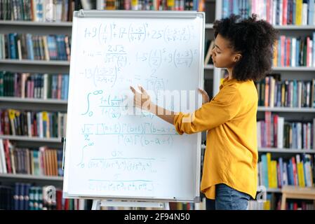 Confident African American intelligent young adult female teacher stand near the whiteboard, conducts lecture at the library, shows information to the students. Distant learning, online conference Stock Photo