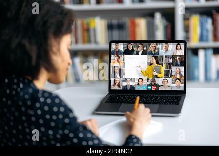 View over shoulder of an mixed race clever female student at a laptop screen with a female teacher and students. Teacher conducts online lecture, female student takes notes. Online training,video call Stock Photo