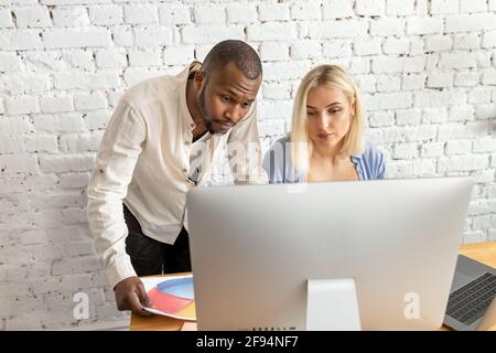 Serious mixed race colleagues using computer, working on report, learning financial graphs. Focused business partners developing new project Stock Photo