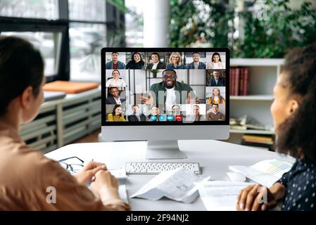 Virtual communication, video call. View over the shoulders of two women to a computer screen with successful multiracial business team gathered in a video conference to discuss working issues,strategy Stock Photo