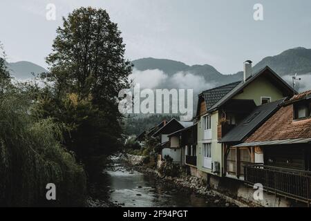 Mountain river and row of houses near it in Slovenian town Bohinjska Bistrica under the Julian Alps hiding in clouds in the background Stock Photo