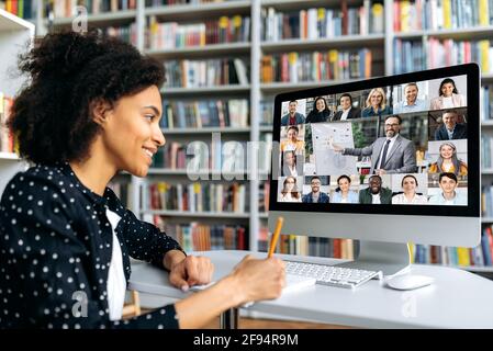 Video conferencing using app and computer. Mixed race female student, studying remotely, listening to an online lecture, taking notes, multiracial smiling people on the computer screen. Video call Stock Photo