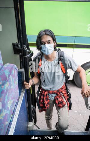 Asian young man upwards wearing face mask and backpack about to take the bus at the bus station. Stock Photo