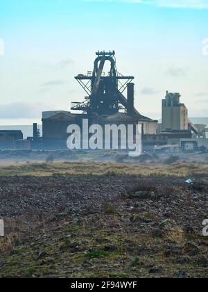 The derelict, abandoned hulk of the blast furnace at the Redcar Steelworks in a haze against a light blue sky, seen from the rubble of the slagfields Stock Photo