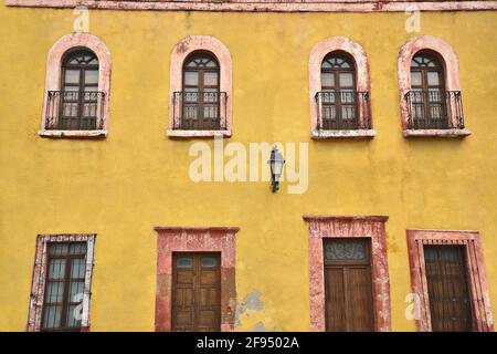 Colonial house facade with a Venetian ochre stucco wall and stone trimmed arched windows with handcrafted railing in Santiago de Querétaro, Mexico. Stock Photo