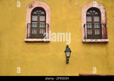 Colonial house facade with a Venetian ochre stucco wall and stone trimmed arched windows with handcrafted railing in Santiago de Querétaro, Mexico. Stock Photo