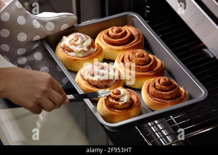 Woman smears finished buns with cream. Cinnamon rolls are baked in the oven.  Homemade baking. Stock Photo