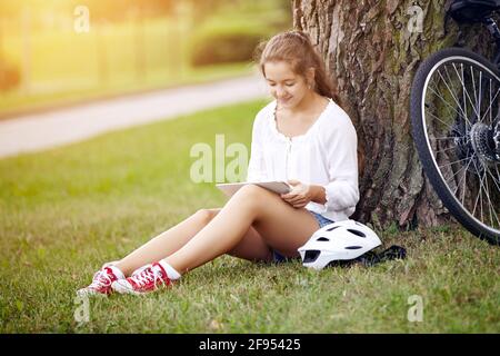 A beautiful young girl sits in a park on the grass and uses a tablet. Technology internet modern lifestyle concept. Stock Photo