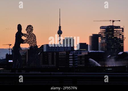 17 March 2021, Berlin: The sculpture 'Molecule Man' (l-r), the Berlin TV Tower and a new building form a panorama in the evening after sunset. The three-part monumental work stands in the Spree between Elsenbrücke and Oberbaumbrücke (rear), near the intersection of the three city districts of Kreuzberg, Treptow and Friedrichshain. Photo: Stefan Jaitner/dpa Stock Photo